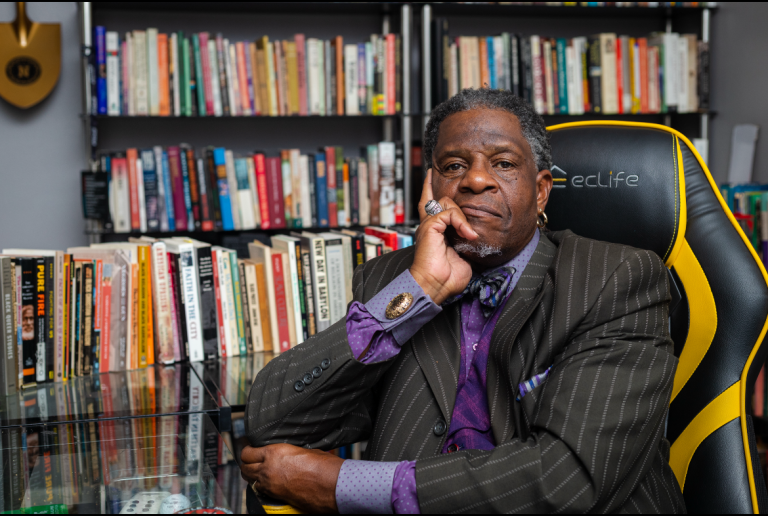 A man with his hand resting on his chin, dressed in a suit and surrounded by a bookshelf filled with books, appears to be indoors, likely in an office.